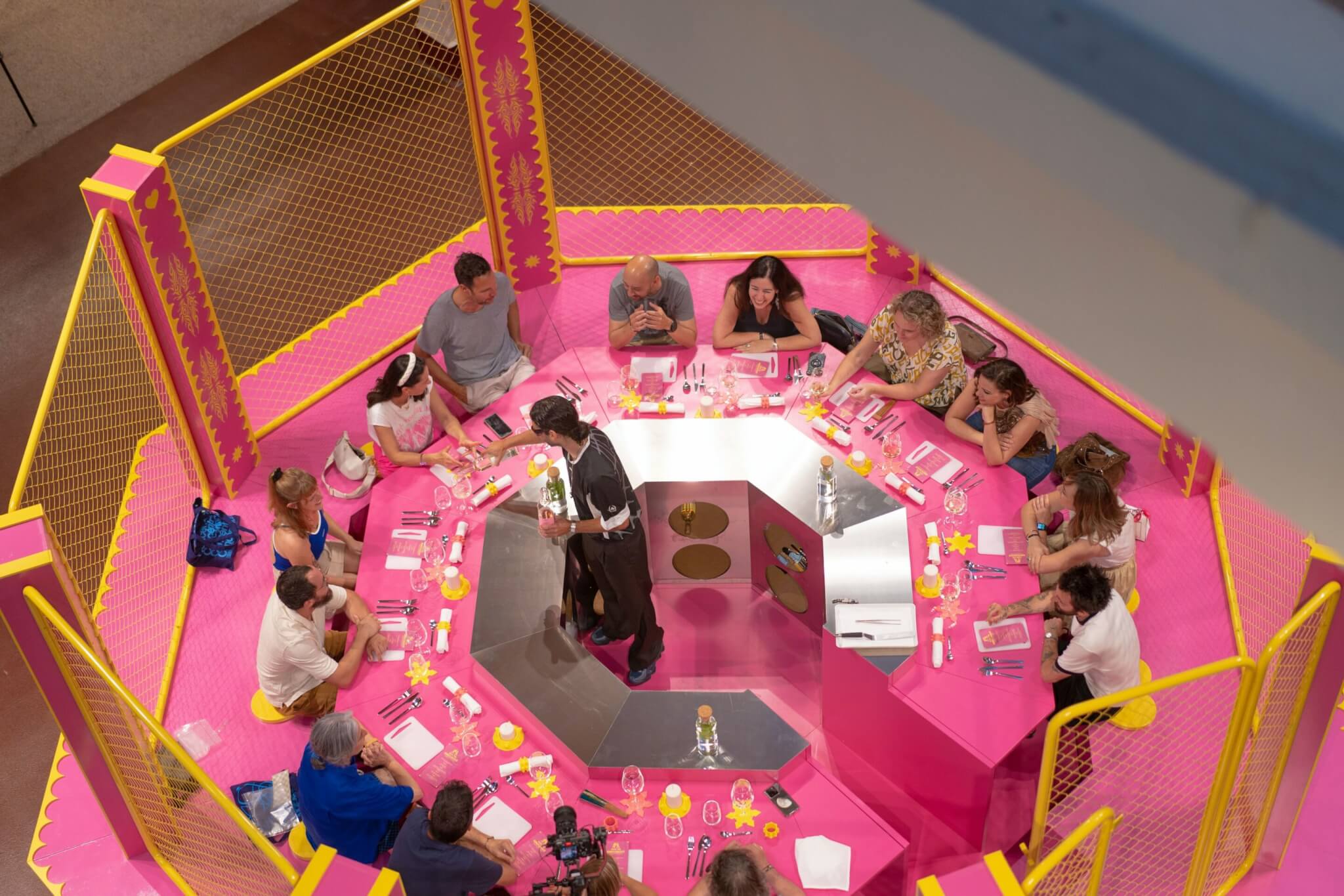 pink art installation shaped like an MMA ring. In the center a man serves food to patrons sitting around the perimeter. 