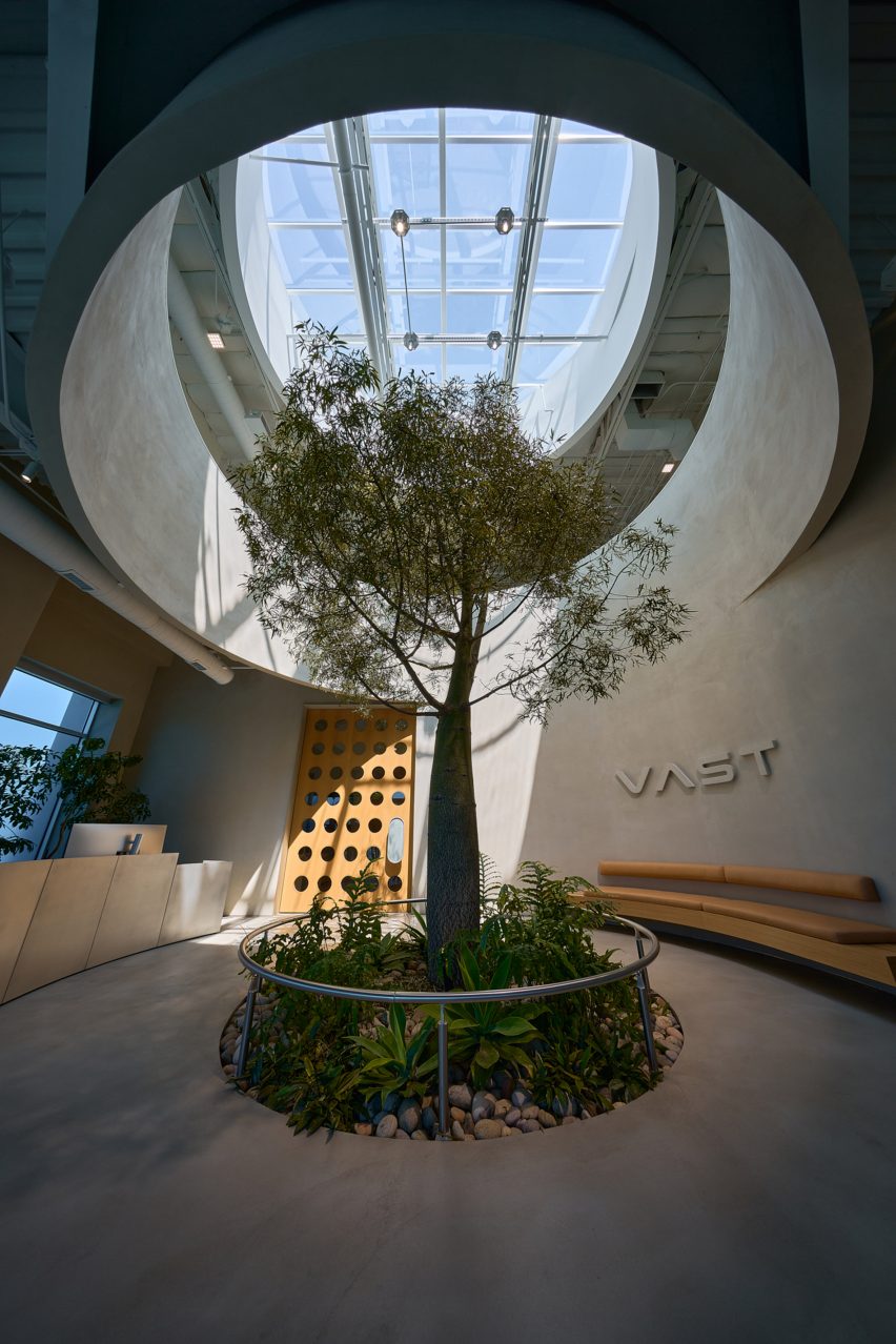Photo of the lobby of Vast's headquarters in Long Beach, showing a room with chamfered corners, a tall tree planted in the ground, a circular oculus cut into the mezzanine above and a circular skylight beyond that