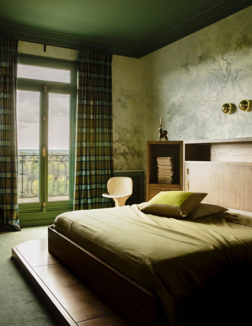 A photograph showing the master bedroom, with a green colour scheme. A large double bed sits on a wooden platform and has a wooden headboard. Two tall French windows with green tartan curtains lead out to a small balcony.