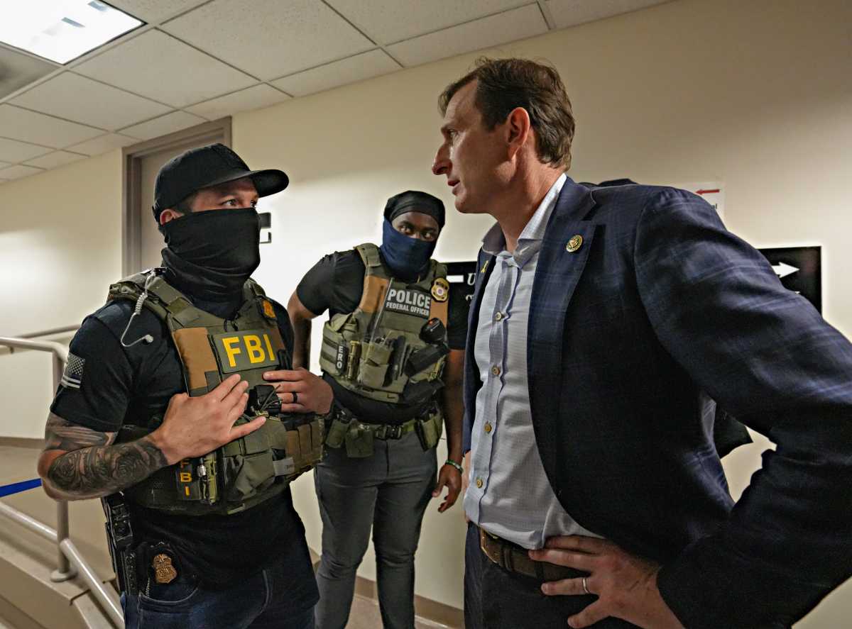 Man in mask looks back at man in suit, with latter providing a stern gaze and hands on hips