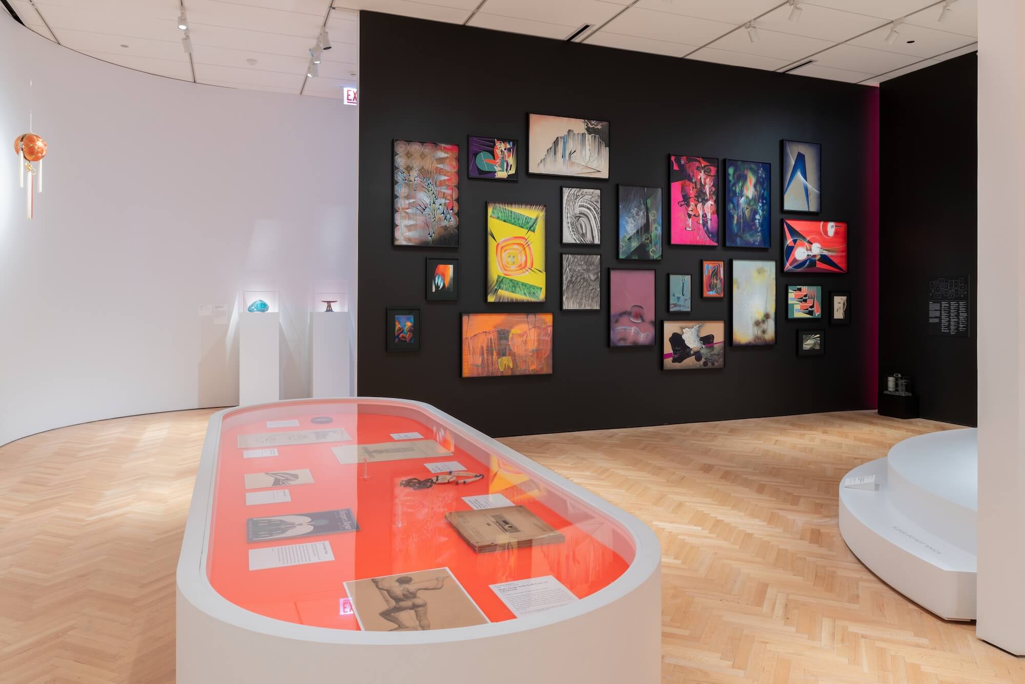 A view of the exhibition Bruce Goff: Material Worlds featuring an oval vitrine and a black gallery wall featuring several abstract paintings.