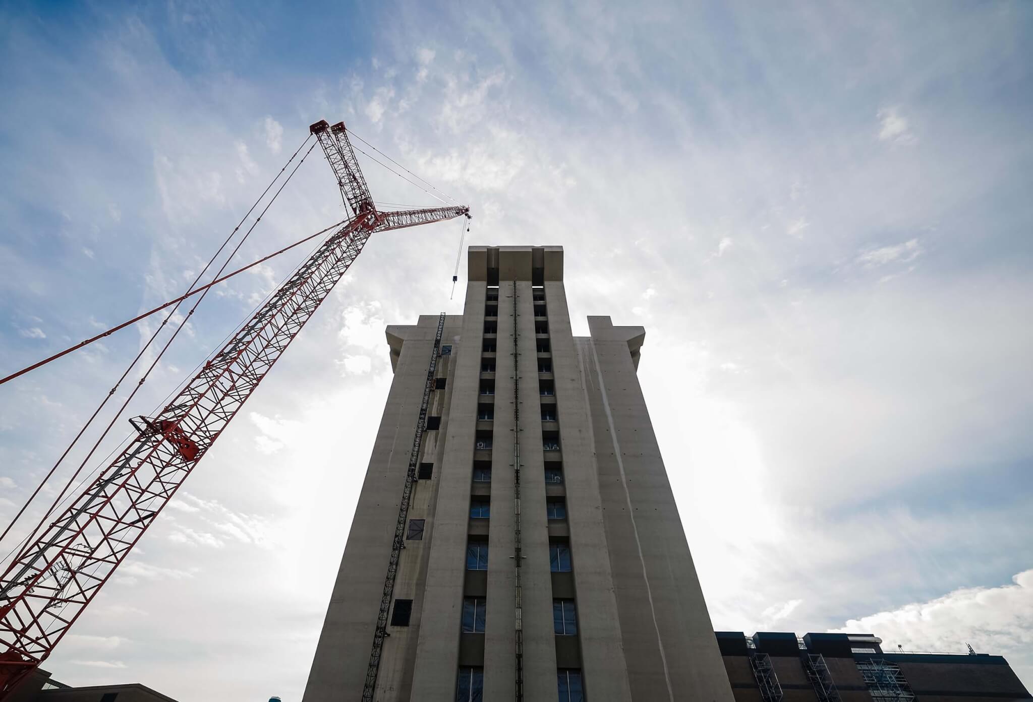 Low-angle view looking up at Crosley Tower during demolition, with a red crane extending to the left and the tower's concrete facade and flared crown rising against a cloudy sky.