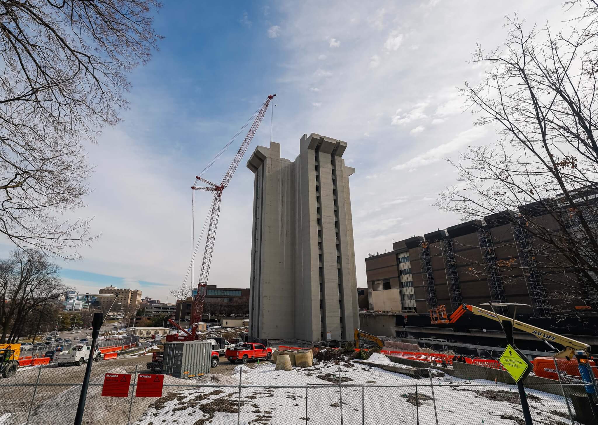 Crosley Tower mid-demolition seen through a chain-link fence in winter, with snow on the ground, construction vehicles and orange barriers in the foreground, and adjacent campus buildings visible at right.