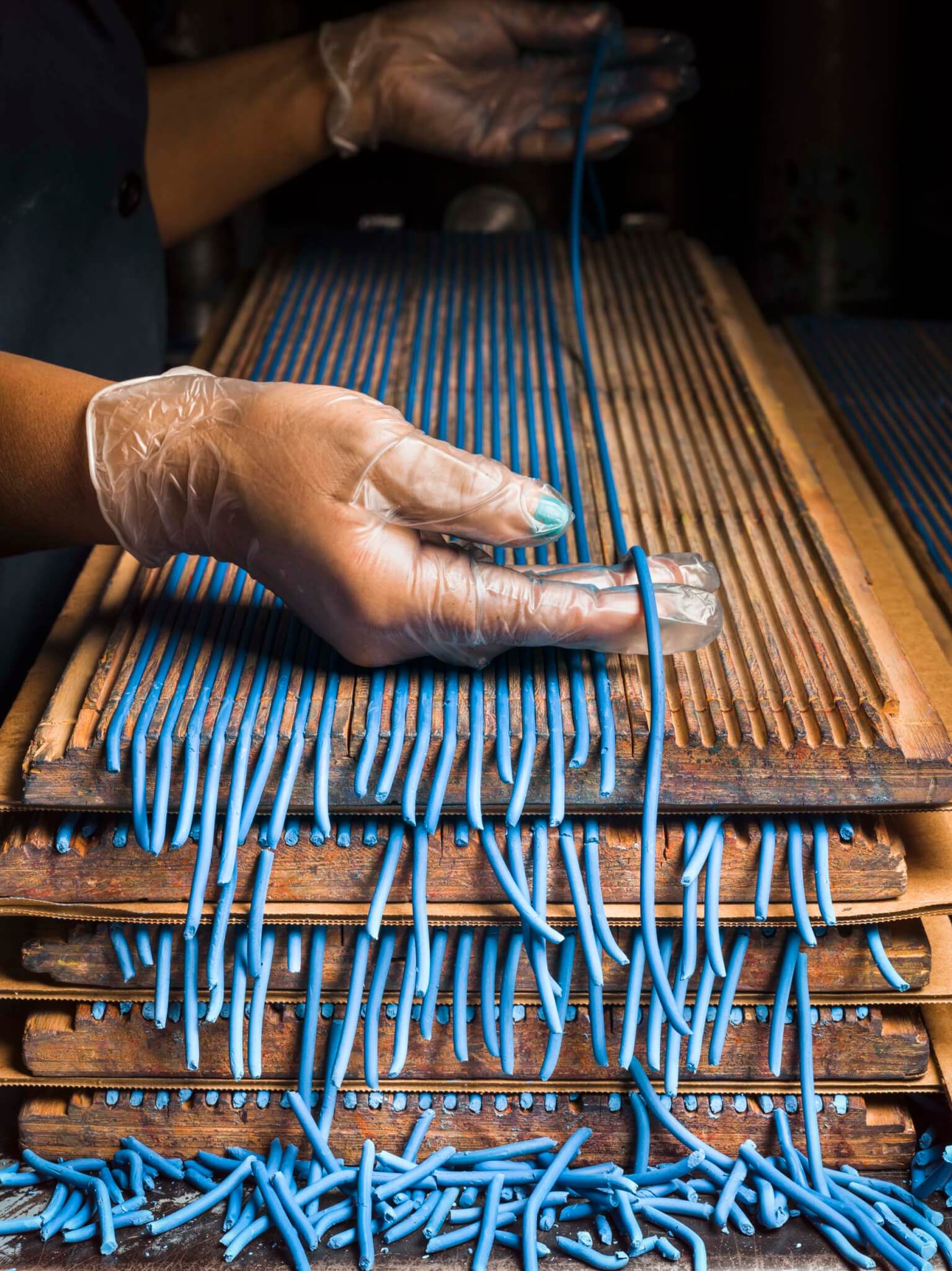 A gloved hand sorts freshly extruded blue pencil cores draped over wooden drying racks, with trimmed pieces scattered below.