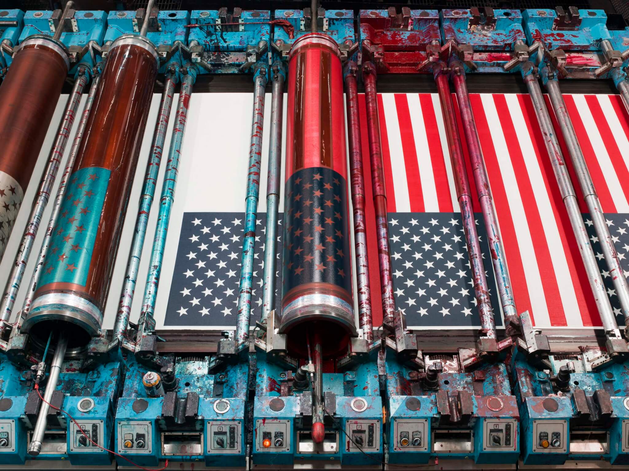 A top-down view of an industrial printing press with ink-splattered blue rollers producing American flags.
