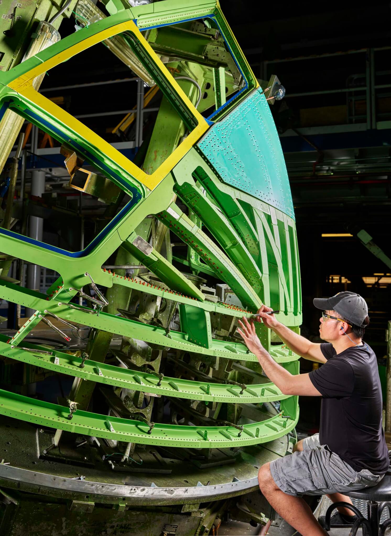  A worker in a black cap kneels to inspect the green-painted structural ribs and skin panels of an airplane nose cone
