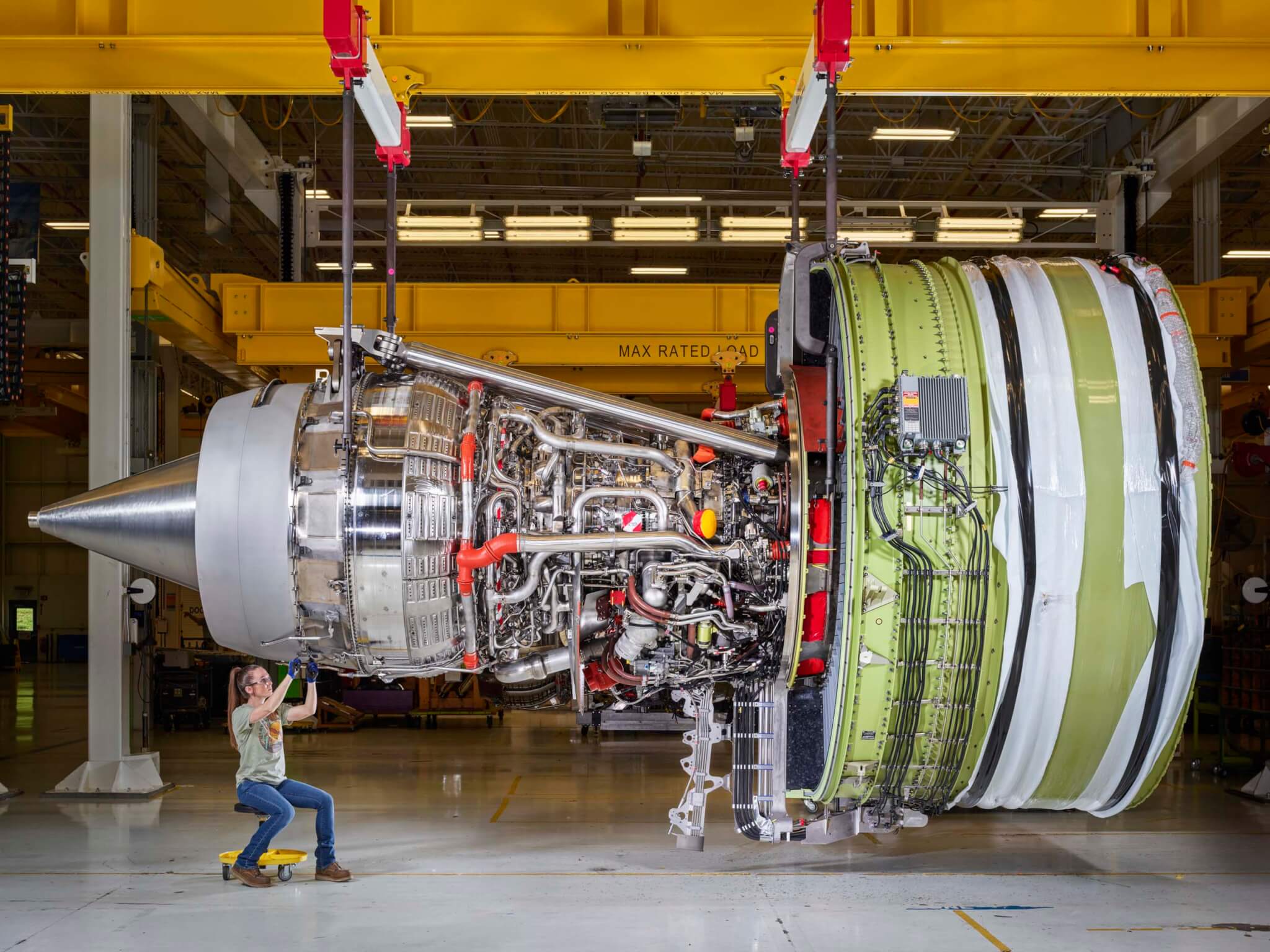 A technician seated on a rolling stool inspects a massive exposed jet engine