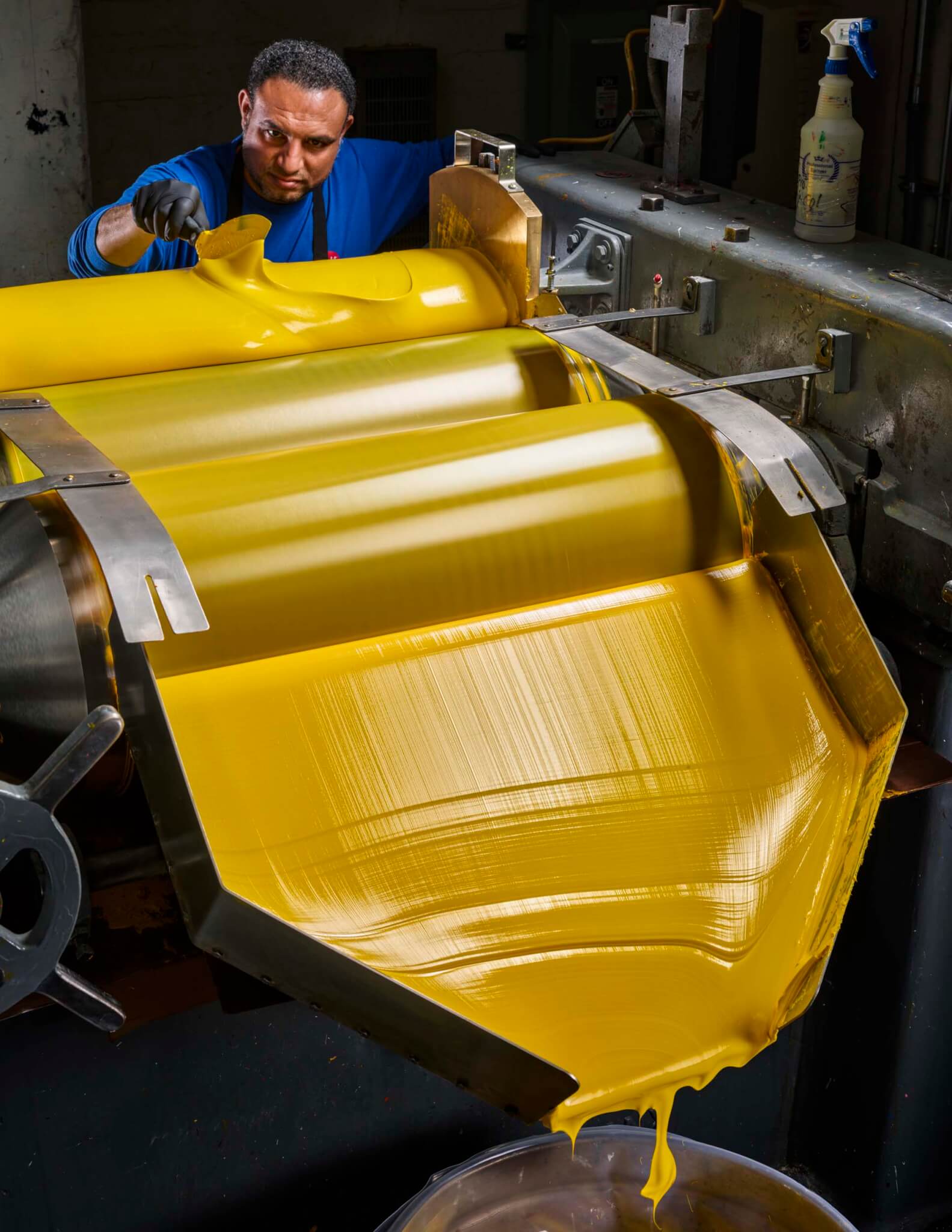 A man in a blue shirt and black gloves leans over a three-roll mill as thick yellow ink cascades