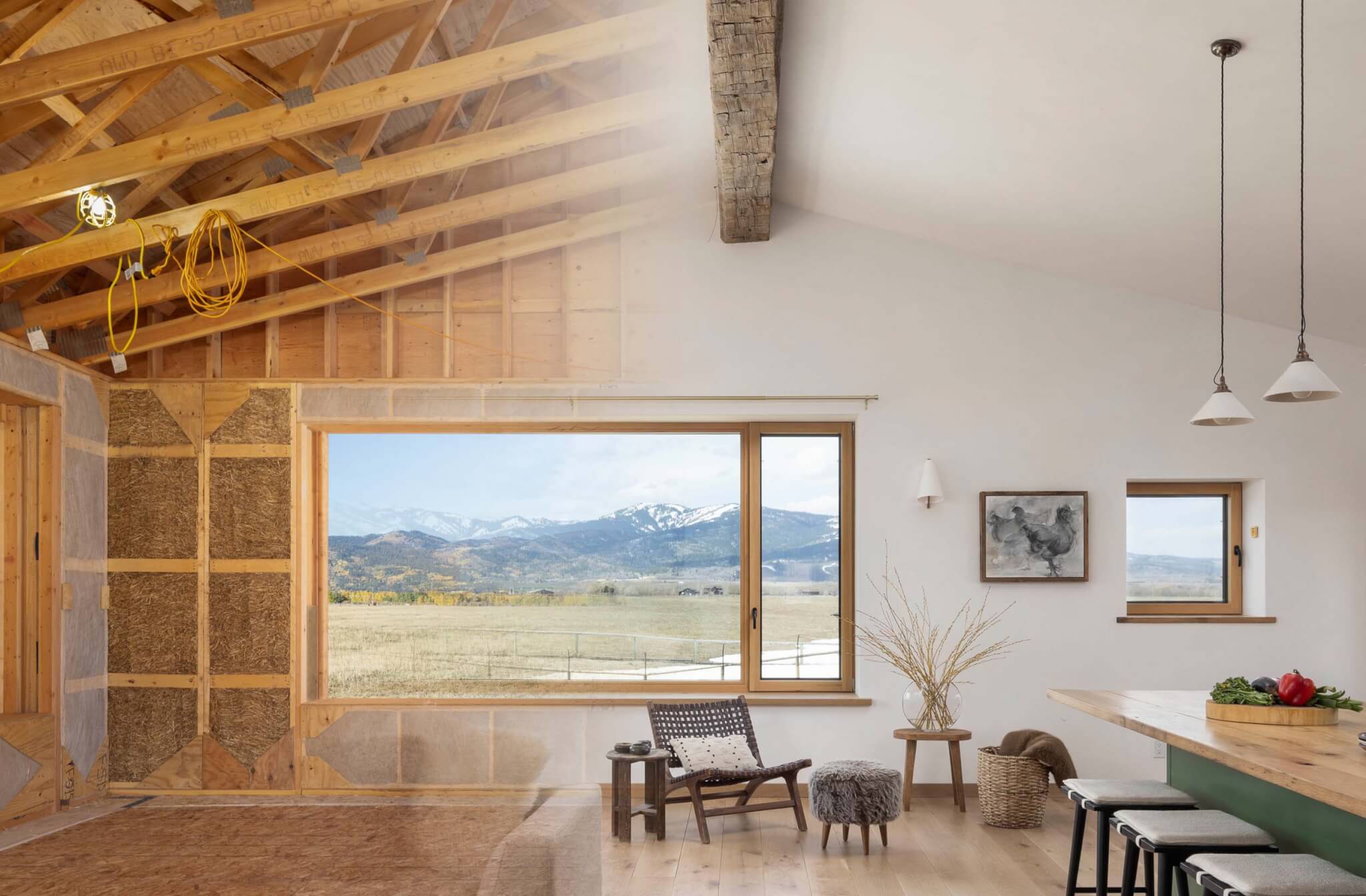 Side-by-side composite image showing the same room during construction with exposed straw panels and wood trusses, and after completion with white plaster walls, wood flooring, a large picture window framing mountain views, and a furnished living and dining area
