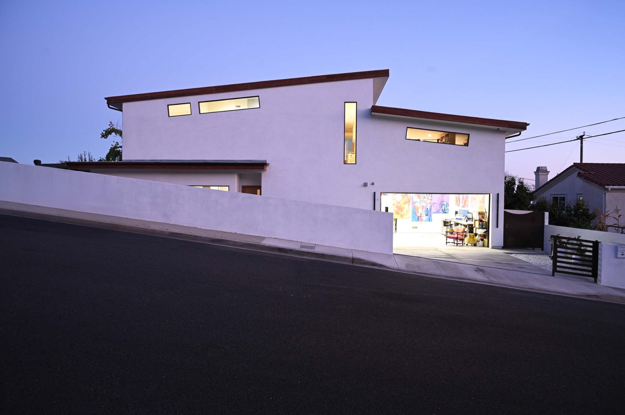 A two-story white stucco house photographed at dusk from the street, with a sloped butterfly roof, narrow horizontal clerestory windows, and an open garage door revealing a brightly lit art studio with large colorful paintings inside.