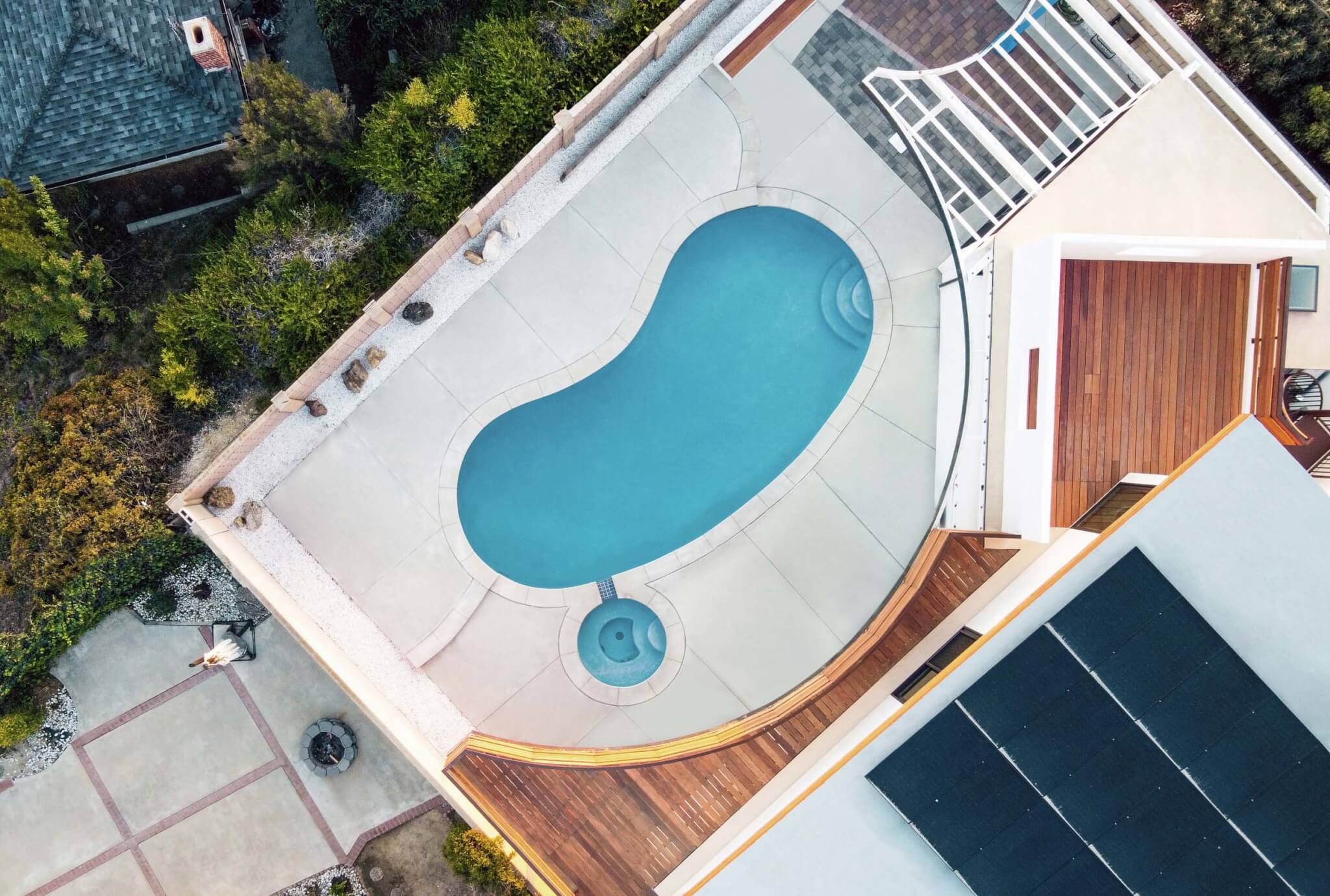 Overhead aerial view of a kidney-shaped swimming pool with an adjacent hot tub, embraced by a curving teak wood deck with solar panels visible on the adjacent roof.