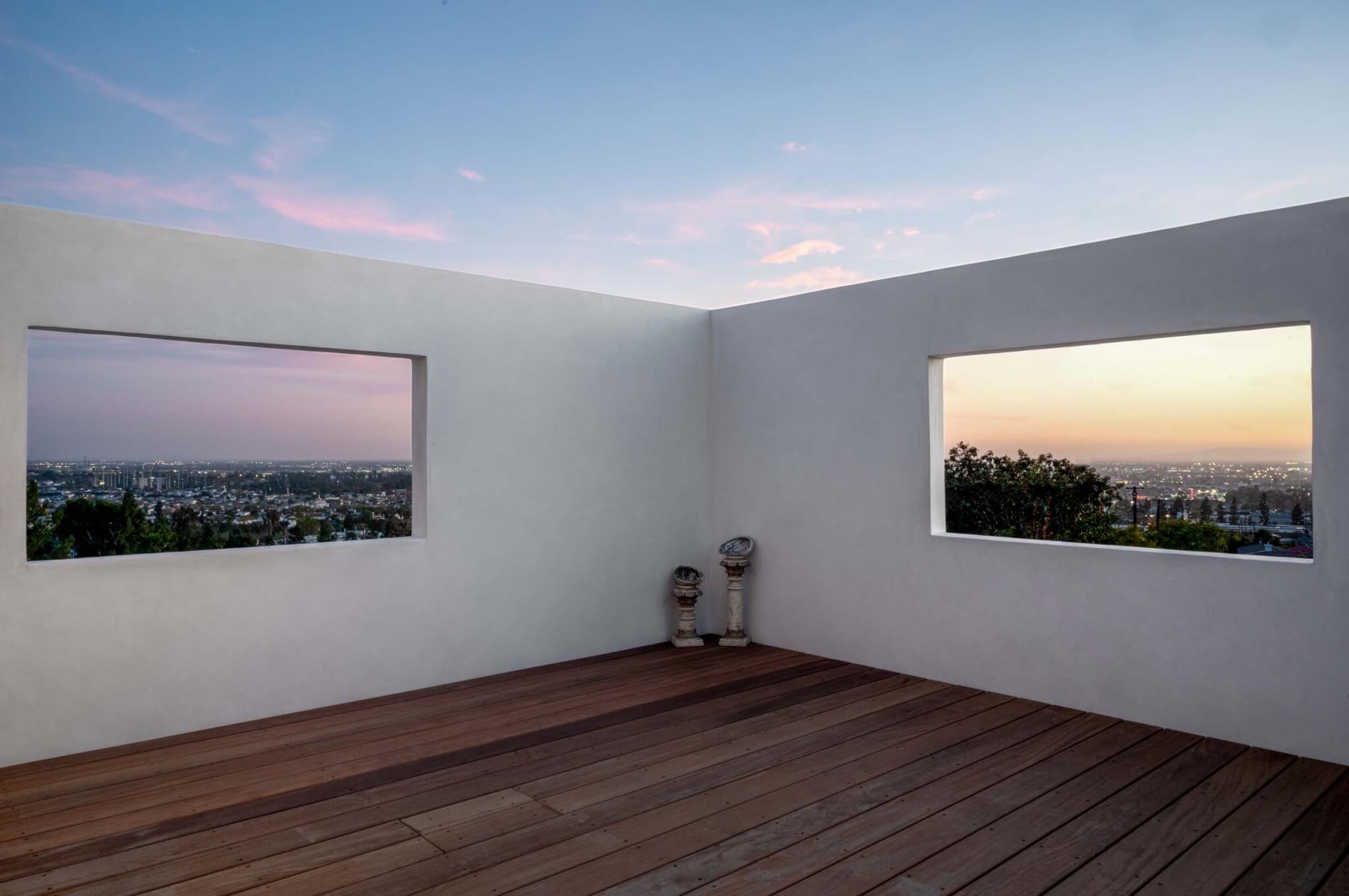 An outdoor teak deck at sunset enclosed by white stucco walls with two large rectangular window openings showing panoramic views of the Los Angeles basin in pink and blue twilight tones, with a small decorative sculpture in the corner.